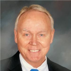 Professional business headshot of Roger Minkle, middle-aged man with light hair wearing dark business suit and blue tie, smiling confidently at camera